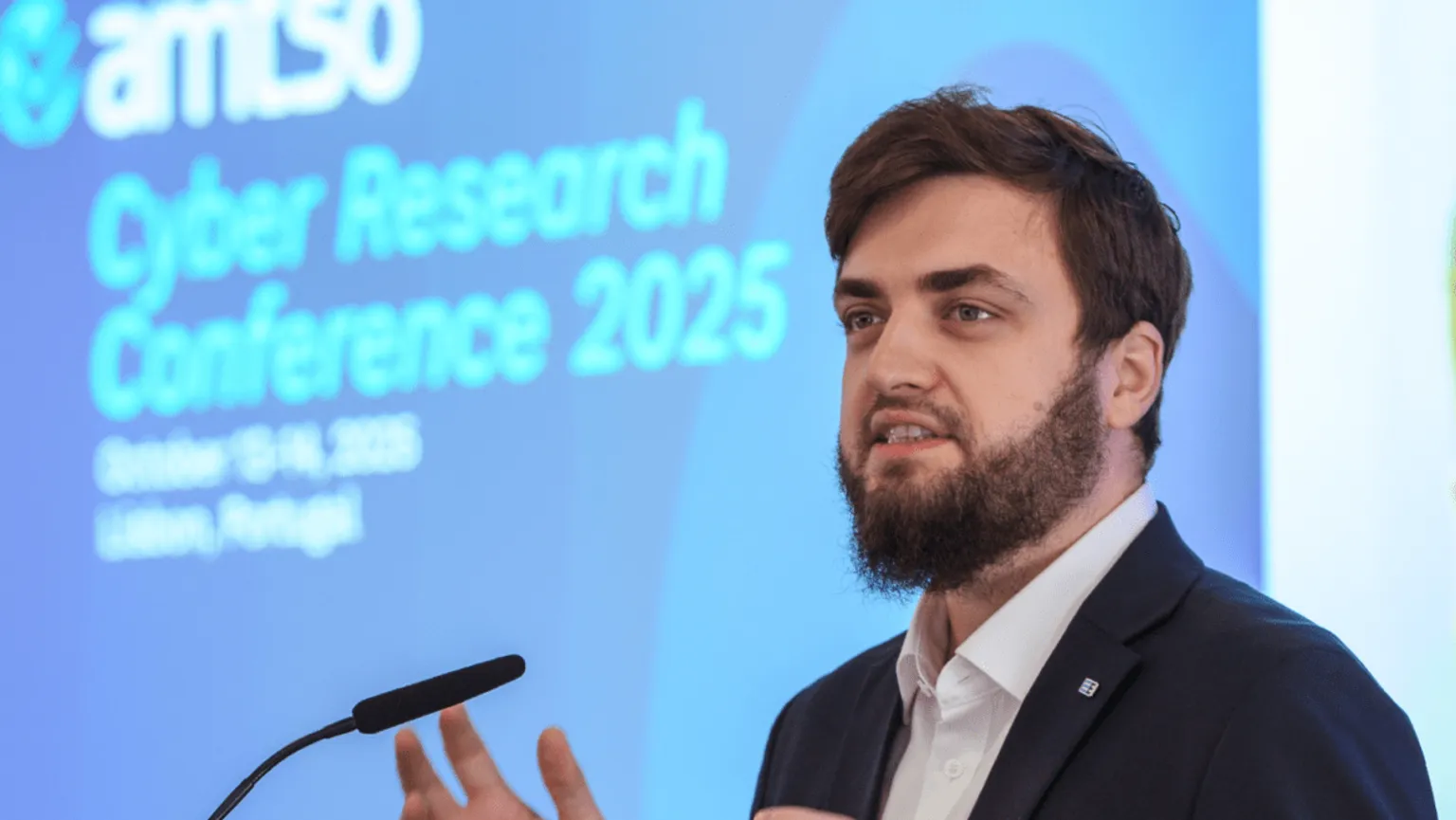 Vlad Iliushin, ELLIO Founder and AMTSO President presenting at cybersecurity conference with "Cyber Research Conference 2025" displayed on blue projection screen behind podium