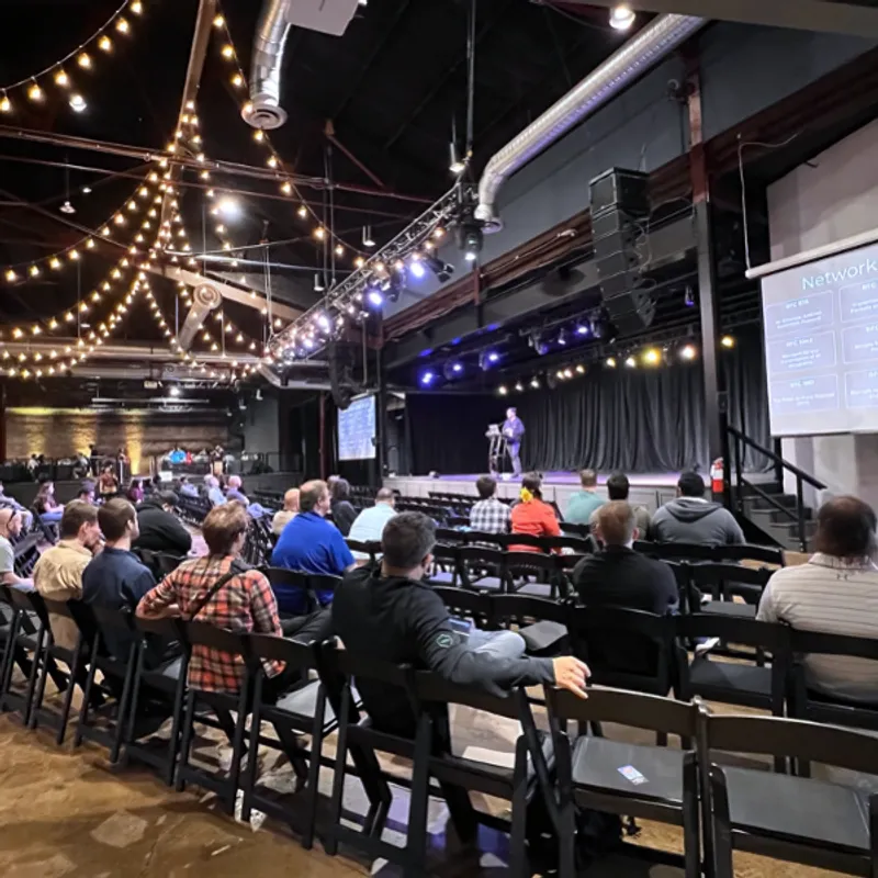 Conference presentation in industrial venue with audience seated at tables, speaker on stage with projection screens displaying cybersecurity content