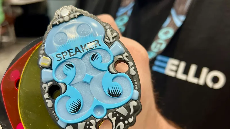 Blue speaker DEF CON 33 badge with decorative bone details, held in hand with colorful lanyard, ELLIO branding visible in background