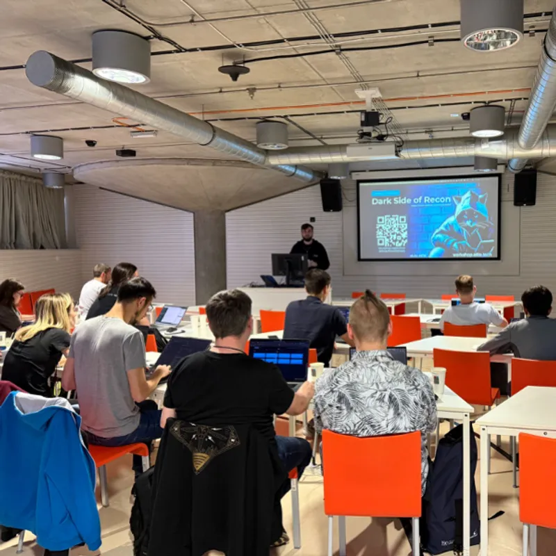 Cybersecurity presentation in modern conference room with attendees at orange chairs viewing "Dark Side of Recon" slide on large screen