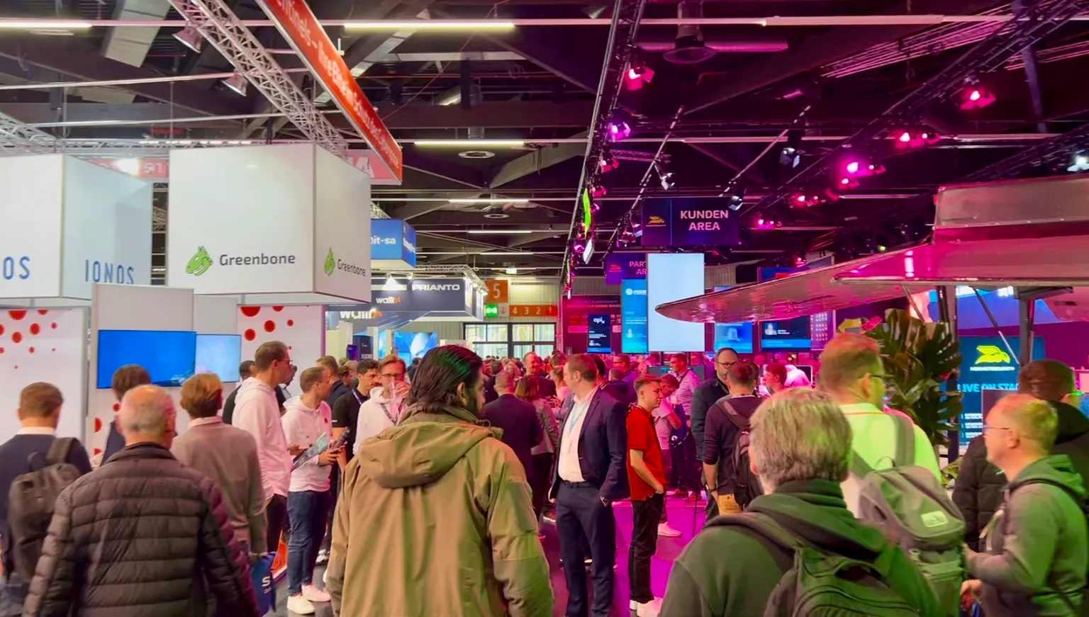 Crowded technology trade show floor with colorful booth displays including Greenbone cybersecurity company, attendees networking under purple lighting and "KUNDEN AREA" signage